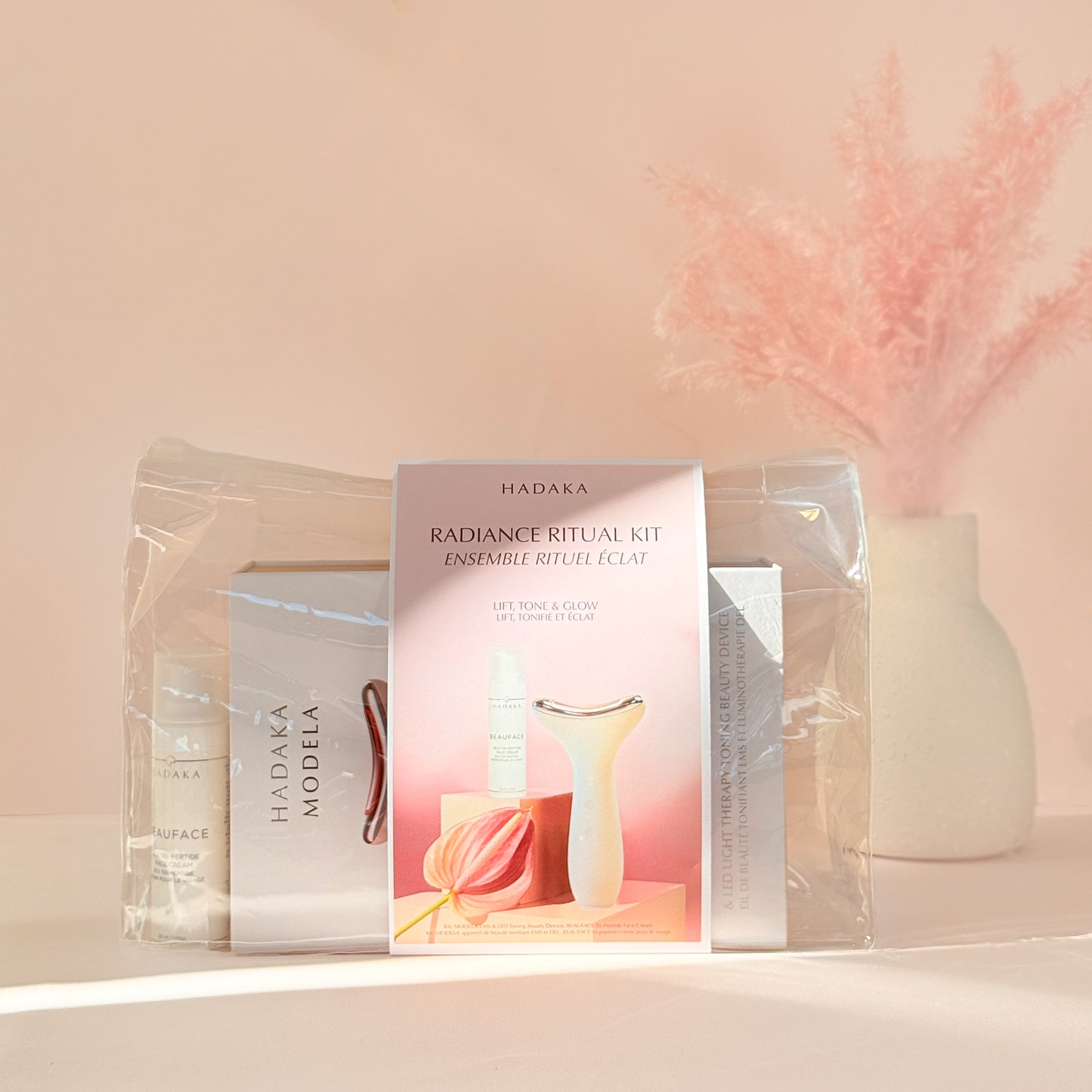 Hadaka Radiance Ritual Kit with MODELA EMS & LED Device and BEAUFACE Tri-Peptide Cream on a light pink background with decorative elements.