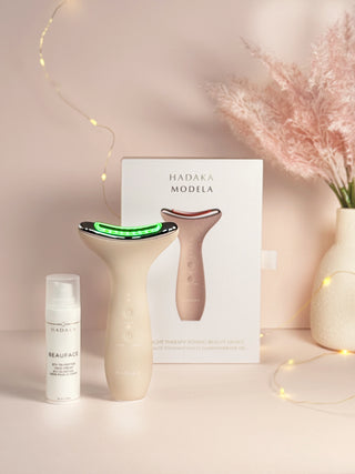 Anti-aging skincare gift set featuring LED light therapy and microcurrent facial toning tools on a light pink background with decorative elements