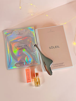 Facial care products by Hadaka including Microcurrent Gua Sha, Beta-Glucan Mask, Marula Oil, and Hair Perfume on a light pink background.